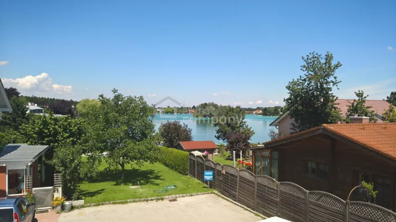 Weitläufiger Blick auf einen See mit umliegender grüner Landschaft und benachbarten Häusern unter blauem Himmel.
