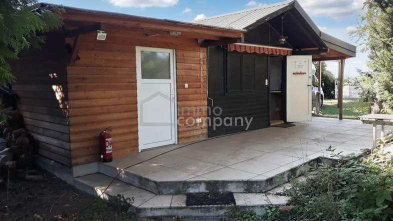 Gepflasterte Terrasse vor einem rustikalen Holzgebäude mit Markise und Blick ins Grüne unter blauem Himmel.