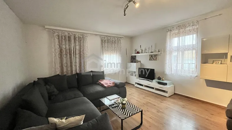 Geräumiges Wohnzimmer mit dunklem Ecksofa, Holzboden und zwei Fenstern, die für natürliches Licht sorgen.