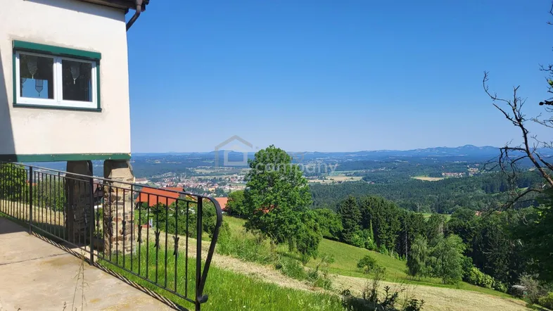 Blick von der Hausseite über eine weite grüne Landschaft mit fernen Ortschaften unter blauem Himmel.