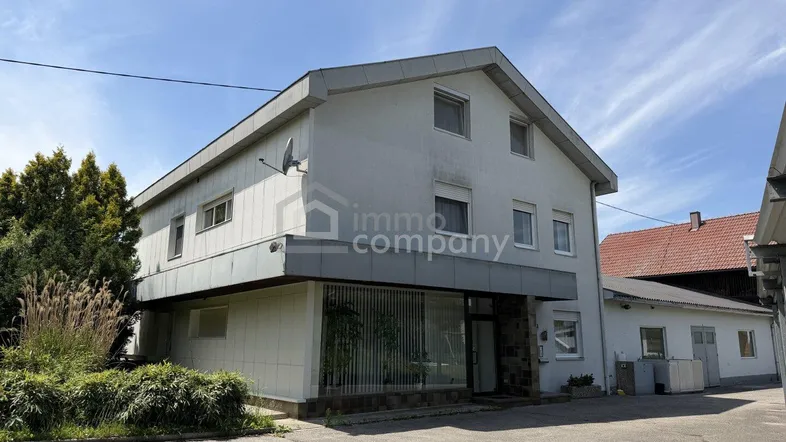 Weißes Geschäftsgebäude mit mehreren Fenstern und einem Vordach, umgeben von Grünflächen.