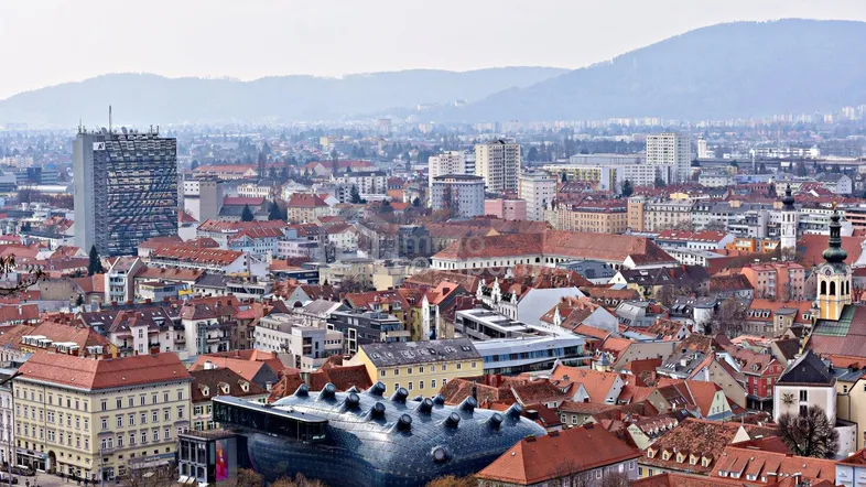 Panoramablick über eine Stadt mit roten Dächern und Bergen im Hintergrund unter bewölktem Himmel.