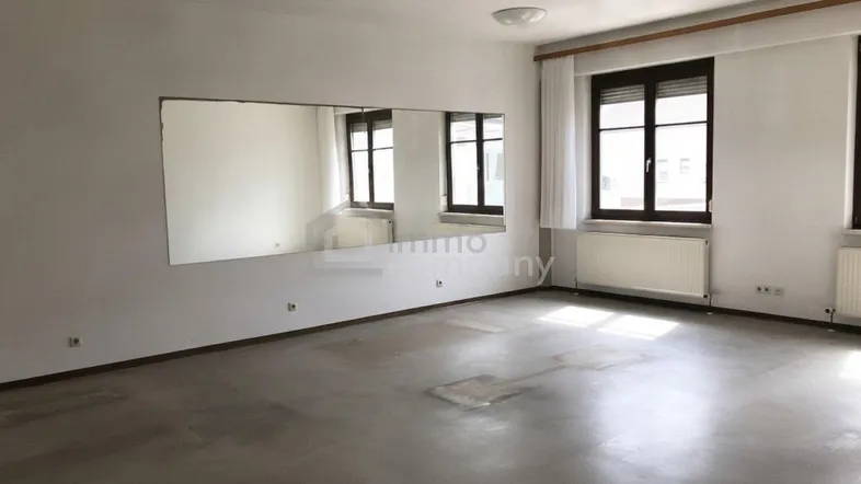 Großer, leerer Raum mit einem Wandspiegel und zwei Fenstern, die Tageslicht hereinlassen.