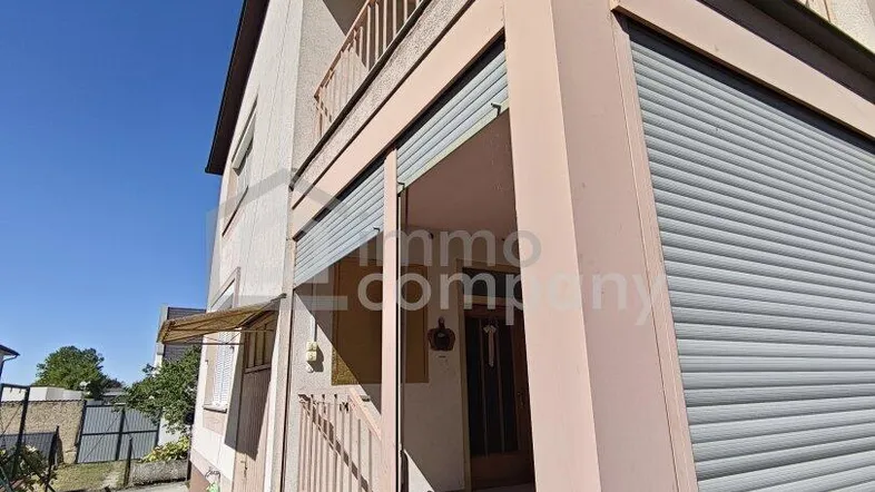 Fassade eines Wohnhauses mit Eingangsbereich, Balkon und Rollläden unter blauem Himmel.