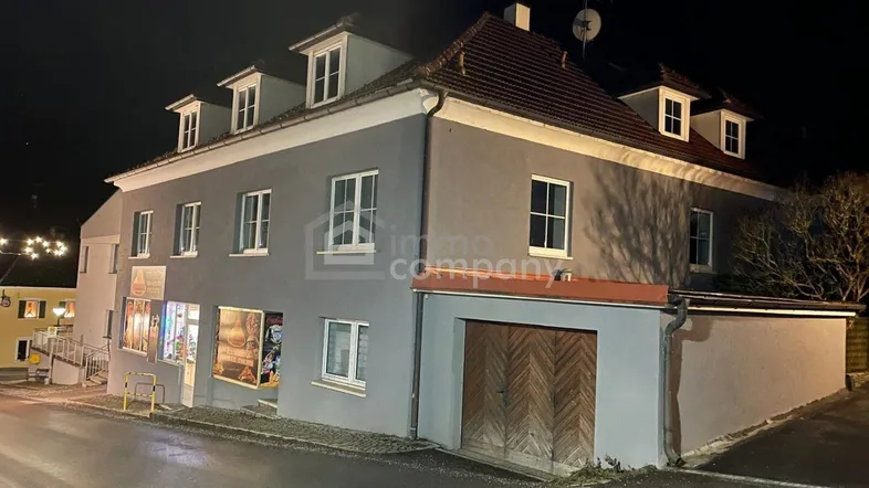 Gebäudeansicht bei Nacht mit grauer Fassade, Dachgauben und einem Geschäft im Erdgeschoss.