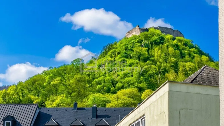 Blick auf eine grüne Hügellandschaft mit einer Burgruine im Hintergrund und Gebäuden im Vordergrund unter blauem Himmel.