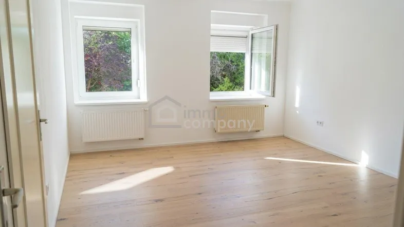 Heller, leerer Raum mit Holzboden, weißen Wänden und zwei Fenstern, die viel Tageslicht hereinlassen.