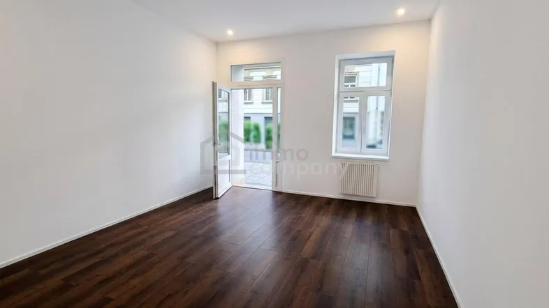 Heller, leerer Raum mit dunklem Holzboden, weißen Wänden und Blick auf die Straße durch Fenster und offene Tür.