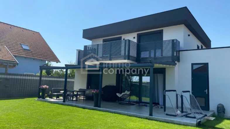 Rückseite des modernen Hauses mit großer Rasenfläche und überdachter Terrasse für Mahlzeiten im Freien.