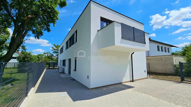 Moderne weiße Fassade eines Neubaus mit klaren Linien und gepflasterter Zufahrt unter blauem Himmel.
