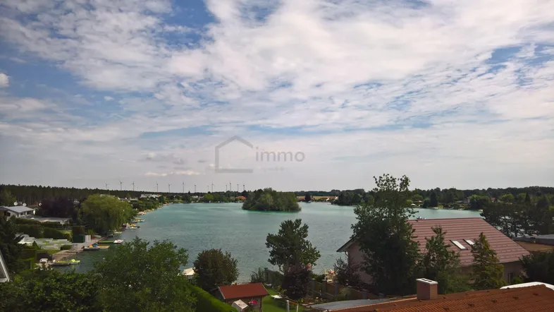 Panoramablick über einen See mit umliegenden Häusern und Windrädern am Horizont unter bewölktem Himmel.