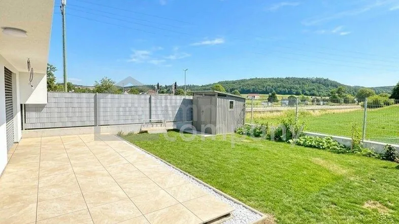 Moderne Terrasse mit Fliesenboden und angrenzendem Rasen, umgeben von grüner Landschaft unter blauem Himmel.