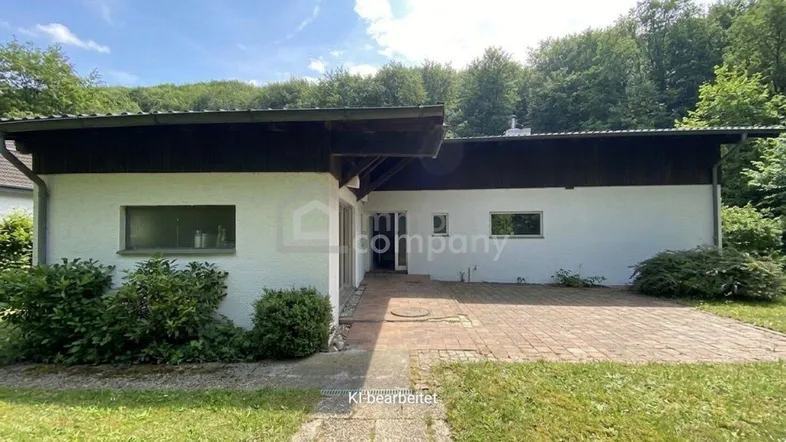 Einfamilienhaus mit weißer Fassade, dunklem Flachdach und gepflastertem Eingangsbereich, umgeben von viel Grün.