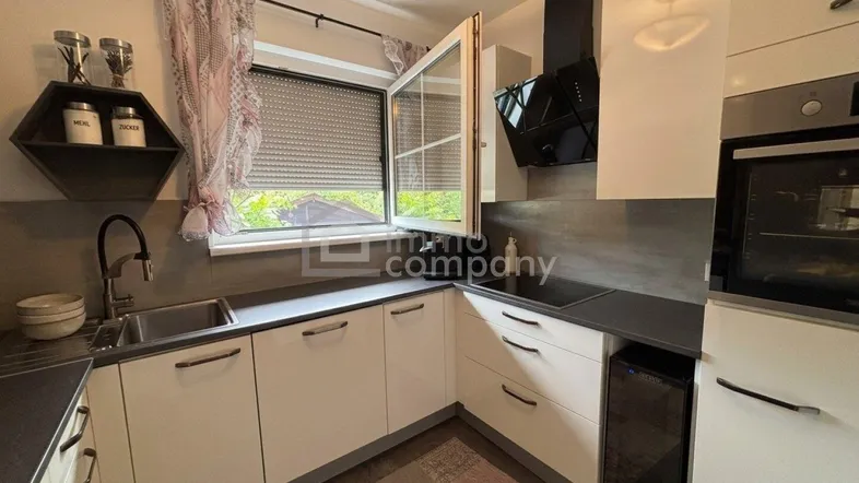 Moderne Küche mit weißen Hochglanzfronten, dunkler Arbeitsplatte und Fenster mit Rollladen.
