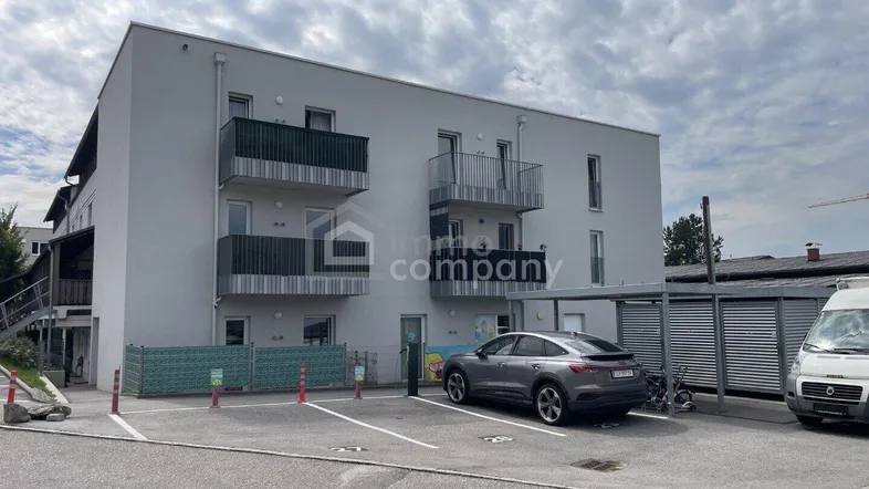 Modernes Mehrfamilienhaus mit Balkonen und Parkplätzen unter bewölktem Himmel.