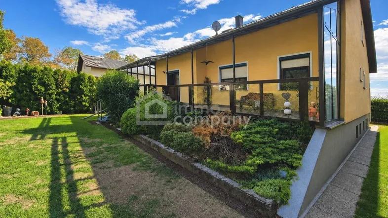 Gelbes Haus mit gepflegtem Garten und überdachter Terrasse mit Glasgeländer.