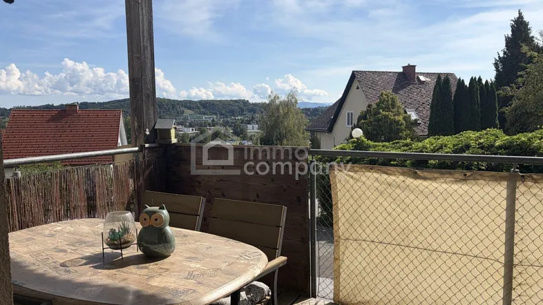 Möblierter Balkon mit Holzboden und weitem Blick über die umliegende Landschaft und Häuser.