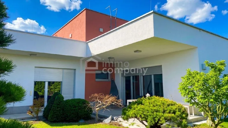 Moderne Hausfassade mit großen Fenstern, Rollläden und gepflegtem Vorgarten unter blauem Himmel.