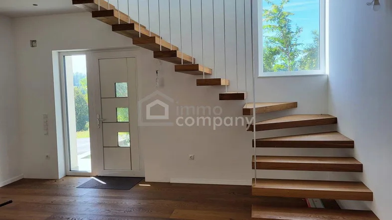 Heller Eingangsbereich mit moderner schwebender Holztreppe und Zugang zum Außenbereich.