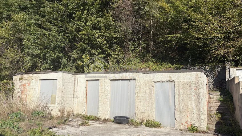 Alte Garagenanlage mit mehreren Toren am Fuße eines bewaldeten Hügels, umgeben von Vegetation.
