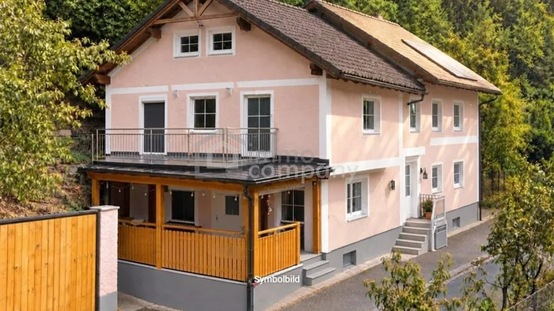 Rosa gestrichenes Einfamilienhaus mit Balkon und überdachter Terrasse, umgeben von viel Grün und Bäumen.