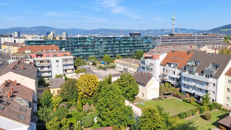 Luftaufnahme einer städtischen Umgebung mit Wohngebäuden, viel Grün und Bergen im Hintergrund.