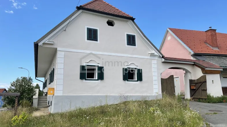 Traditionelles Haus mit Giebeldach und grünen Fensterläden, umgeben von einer Wiese unter blauem Himmel.