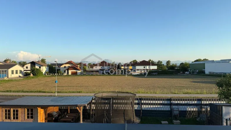 Blick über weite Felder auf Wohnhäuser und ferne Berge unter klarem Himmel.