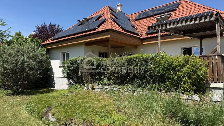 Hausfassade mit rotem Ziegeldach, Solaranlagen und einem grünen Gartenbereich unter blauem Himmel.