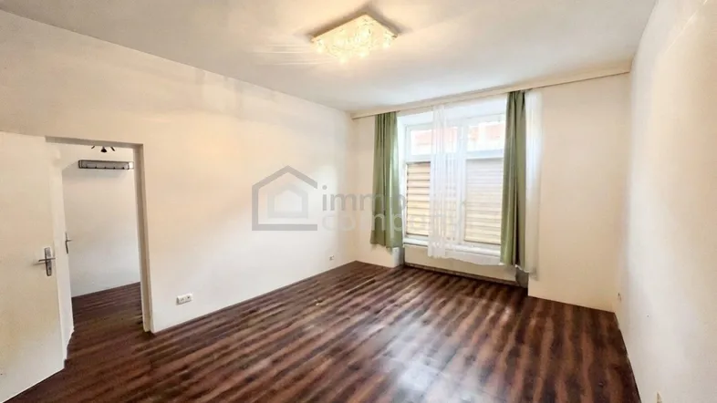 Leerer Raum mit dunklem Laminatboden, weißen Wänden und einem Fenster mit Jalousien.