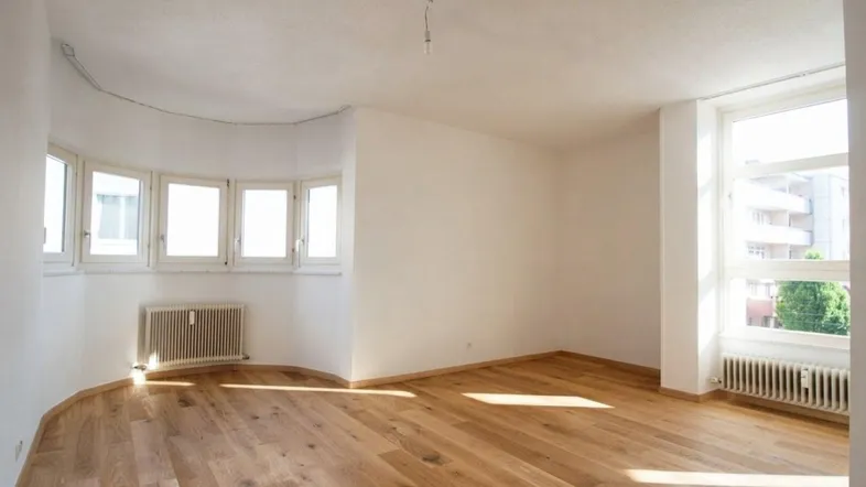 Helles Wohnzimmer mit Holzboden und großen Fenstern, die viel Tageslicht hereinlassen.