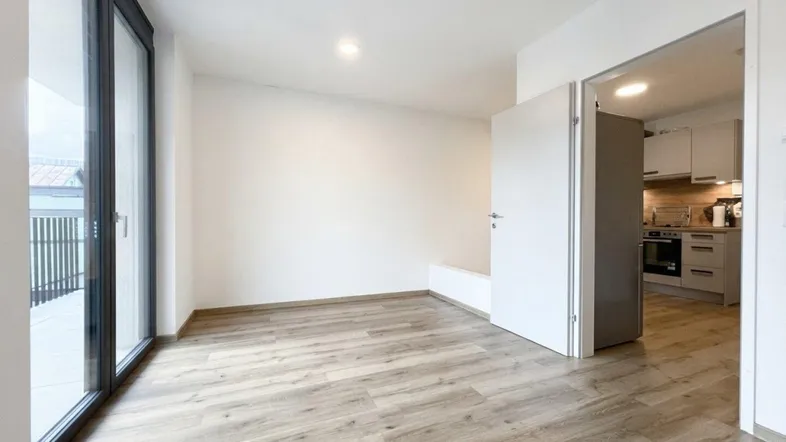 Helles Zimmer mit Holzboden und Zugang zu einem Balkon, angrenzend an eine moderne Küche.