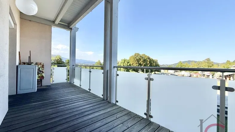 Geräumiger Balkon mit Holzboden, Glasgeländer und weitem Blick auf die umliegende Landschaft und Berge.