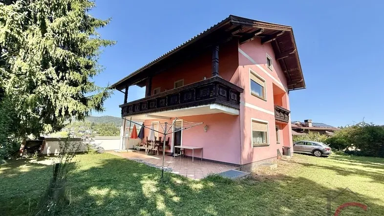 Rosa gestrichenes Haus mit traditionellem Holzbalkon und einer möblierten Terrasse im Gartenbereich.