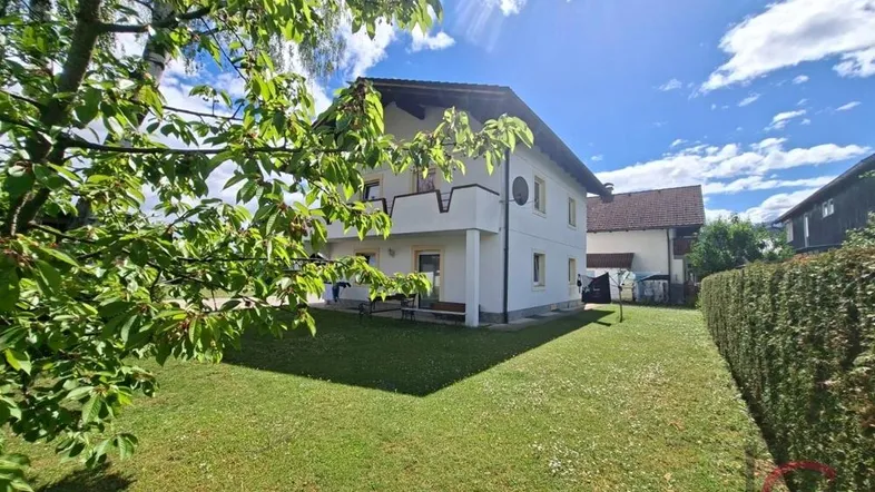 Seitenansicht des Hauses mit gepflegtem Rasen, Bäumen und einer Hecke unter blauem Himmel.