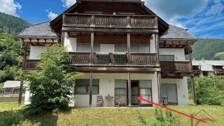 Mehrstöckiges Gebäude im Chalet-Stil mit Holzbalustraden, Terrassenbereich und Bergblick, umgeben von Grünflächen.