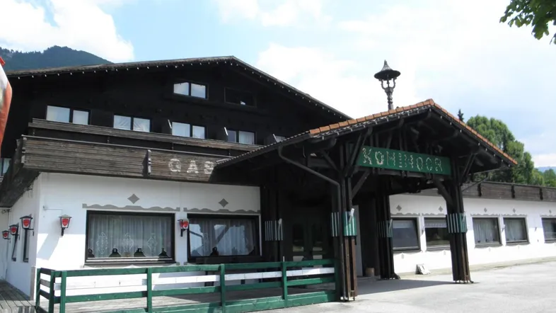 Traditionelles Gebäude mit dunklen Holzbalkonen und weißer Fassade, umgeben von grüner Landschaft.