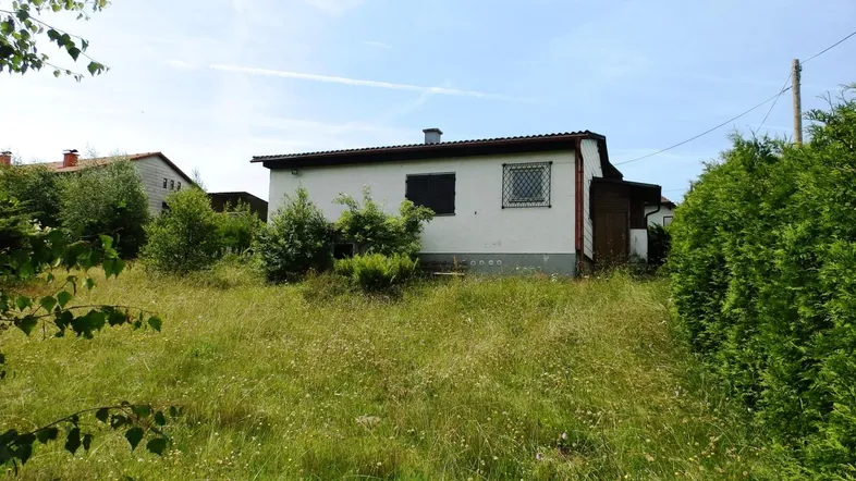 Einfaches Haus mit weißer Fassade und verwildertem Garten unter blauem Himmel.
