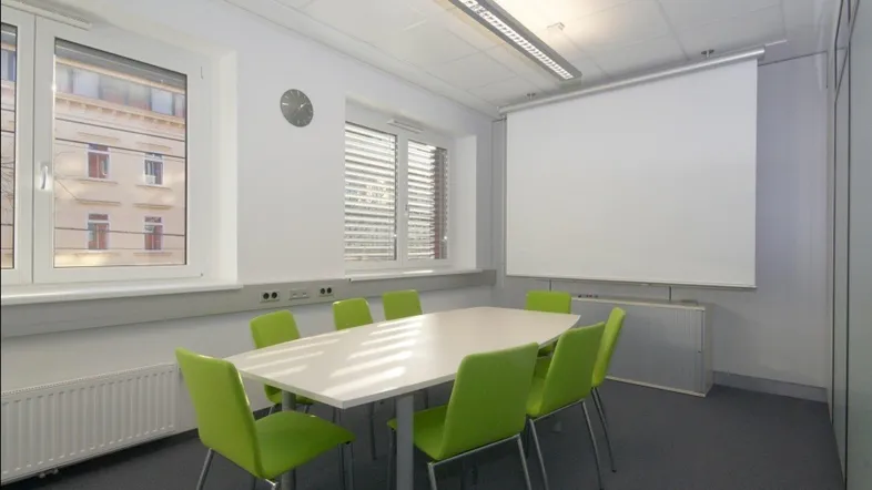 Heller Konferenzraum mit großem Tisch, modernen Stühlen und einem ausziehbaren Projektionsschirm.