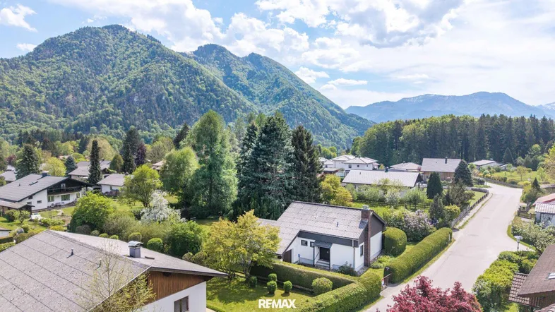 Luftaufnahme einer Wohnsiedlung mit Häusern und üppiger Vegetation vor einer beeindruckenden Bergkulisse.