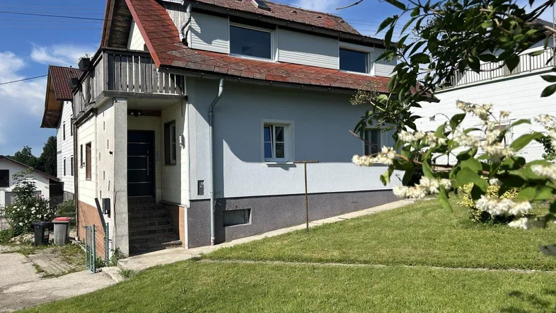 Ansicht des Hauses von vorne mit Gartenbereich und Balkon unter blauem Himmel.