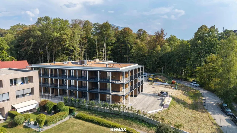 Modernes Gebäude im Bau, umgeben von Bäumen, mit Blick auf die Berge im Hintergrund.