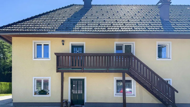 Gelbes Wohnhaus mit Satteldach, Holzbalkon und Außentreppe in ländlicher Umgebung.