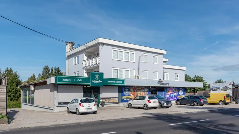 Gebäude mit hellgrauer Fassade und weißen Fensterläden, beherbergt eine Bäckerei, ein Café und eine Spielhalle mit Parkplätzen davor.