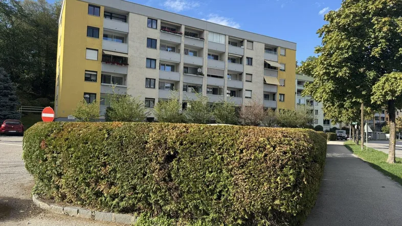 Mehrstöckiges Wohngebäude mit gelber und grauer Fassade, umgeben von Bäumen und Hecken.