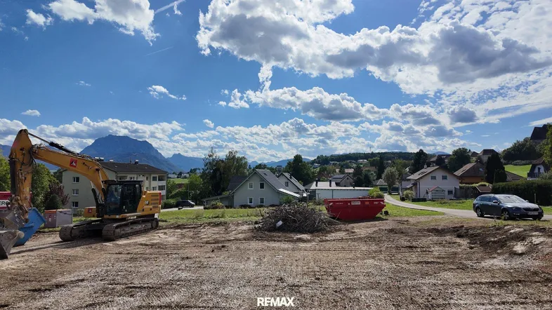 Blick auf eine Baustelle mit Bagger, umgeben von Häusern und einer Bergkulisse unter blauem Himmel.