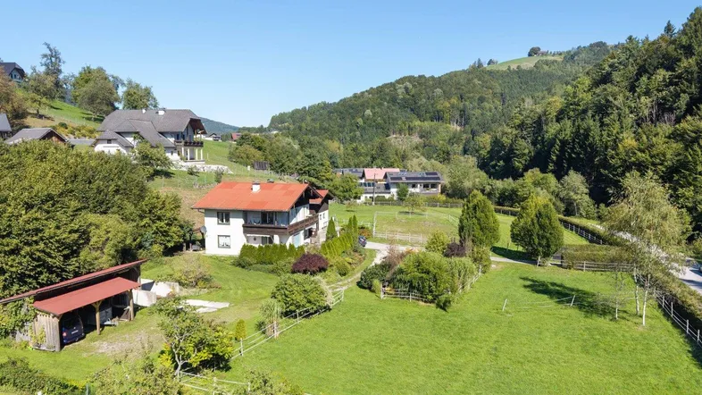 Luftaufnahme eines Anwesens mit mehreren Häusern, weitläufigen Grünflächen und bewaldeten Hügeln im Hintergrund.