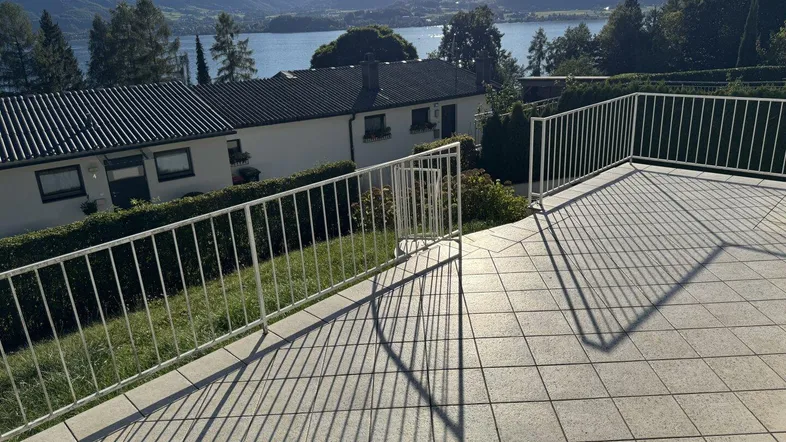 Große geflieste Terrasse mit weißem Geländer und weitem Blick auf den See und die umliegenden Berge.
