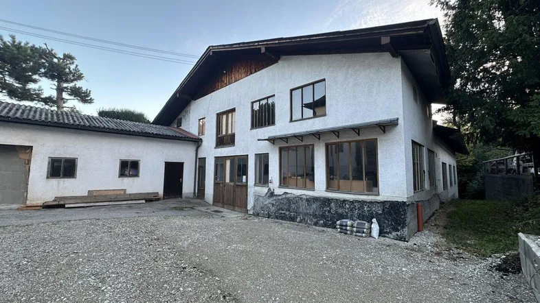 Außenansicht eines Gebäudes mit weißer Fassade, dunklem Dach und angrenzendem Nebengebäude auf einem Schotterplatz.