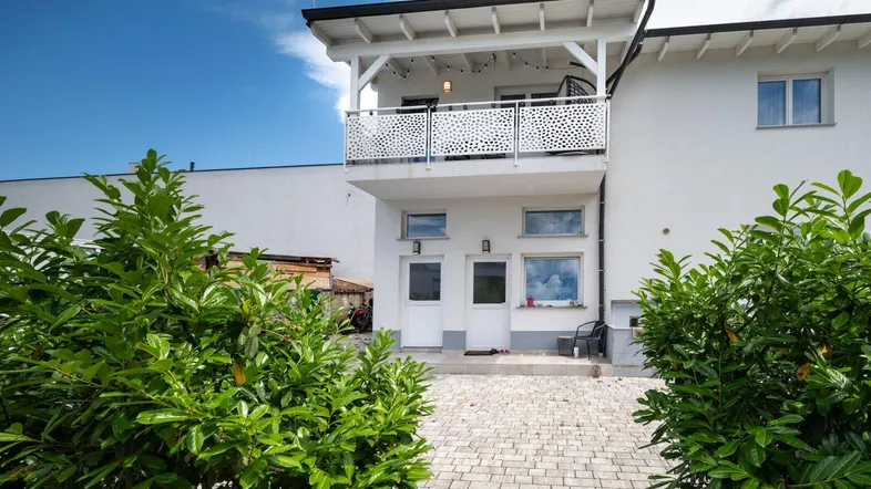 Weißes Haus mit modernem Balkon und gepflastertem Eingangsbereich, umgeben von grünen Büschen.